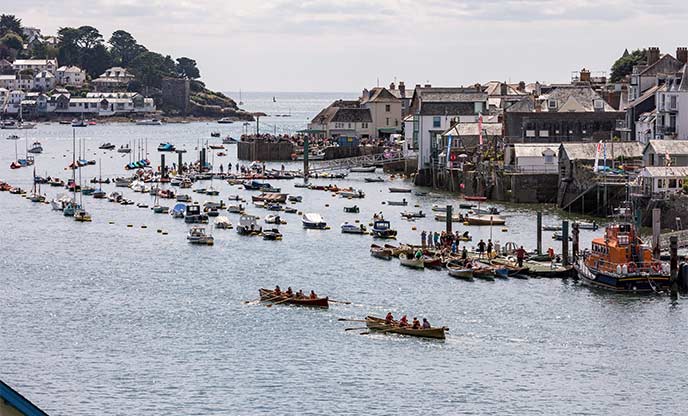 People gig rowing down Fowey River towards boats bobbing in the water