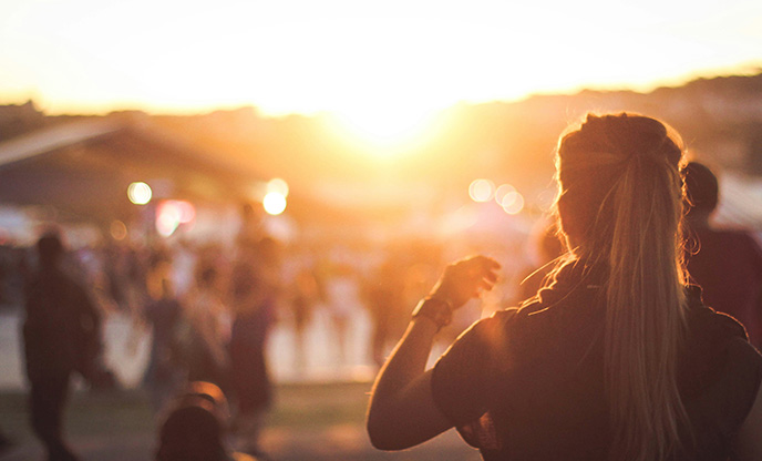 A woman stands in the foreground with her back to the camera and her long hair in a ponytail. In the background the sun is setting, casting everything in a soft light.