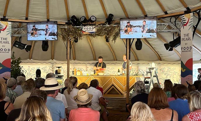 A cooking demonstration is taking place on a stage in a tent, in front of a crowd of people