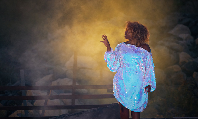 An actress wearing a sequin dress playing the role of Scylla in I AM KEVIN by Wildworks surrounded by rubble and eerie orange smoke