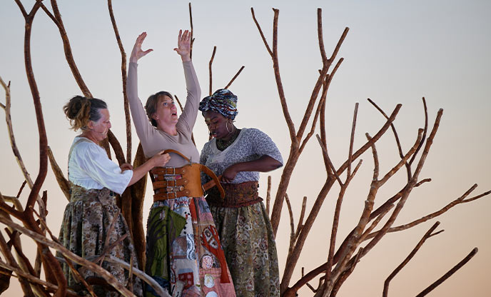 Three actresses standing amidst branches of a tree as dusk during Wildworks' production of I AM KEVIN