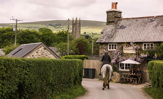 Someone riding a horse down the lane past The Rugglestone Inn, which has flowers climbing up the front