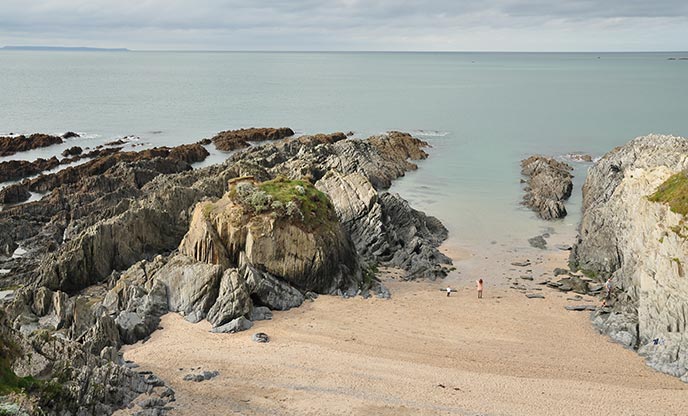 An aerial view of Barrican Beach. There is a relatively narrow stretch of sand between two rocky outcrops, and this gives way to glassy water