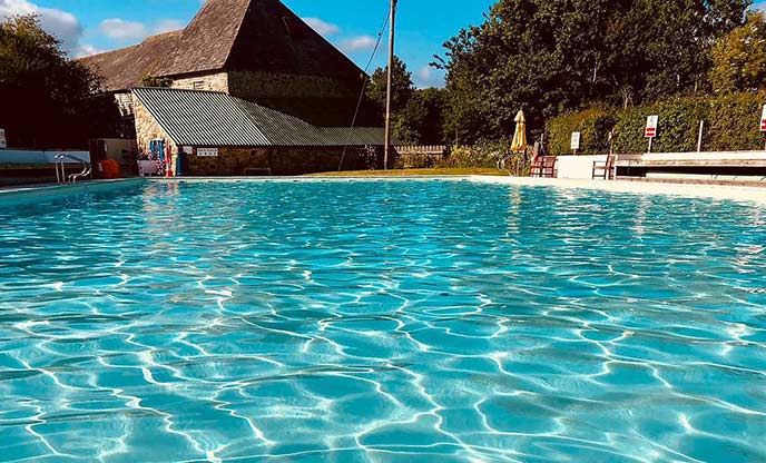 The sun shining through the bright blue water of Chagford Pool, creating patterns on the floor. There is a building in the background..