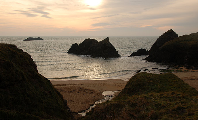 Soar Mill Cove at sunset. The rocks are silhouetted against the sky as they rise from the water.