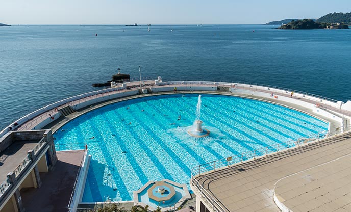 The bright blue water of a lido with a fountain in the centre. The sea visible on the other side of the wall