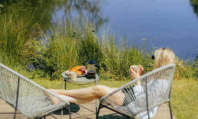Woman sat on a chair beside a lake in the sunshine with a coffee and fresh pastries 