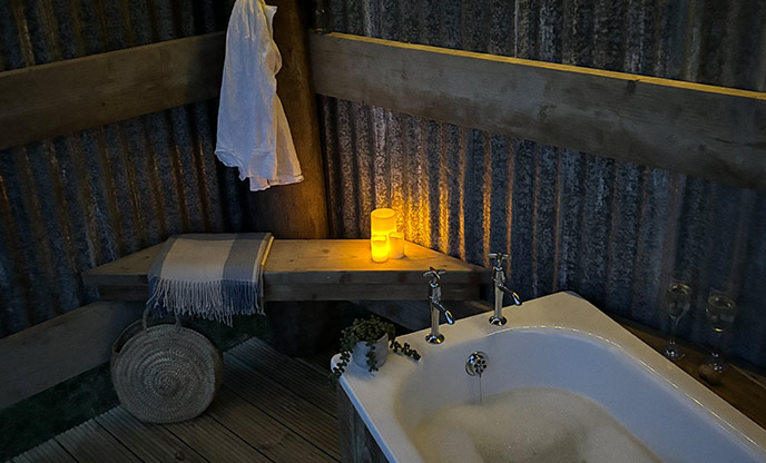 There is a bathtub in the foreground, containing water and bubbles. The wall next to it is made of corrugated steel. There is a bench in the corner with a blanket and a lit candle.