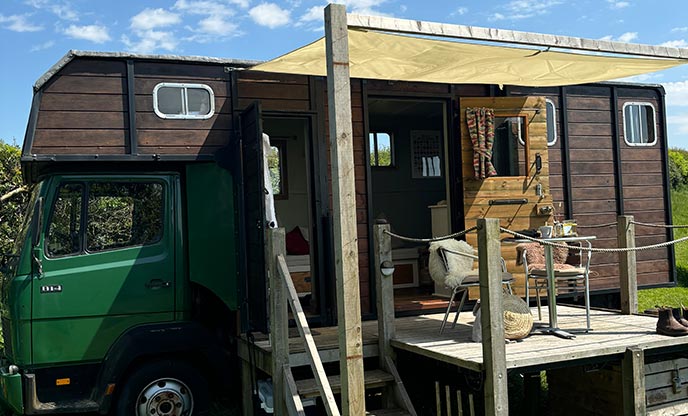 A view of Polly the Lorry, a horsebox which has been converted into accomodation. There is a raised deck outside with a table and chairs on it, shaded from the sun by an awning.