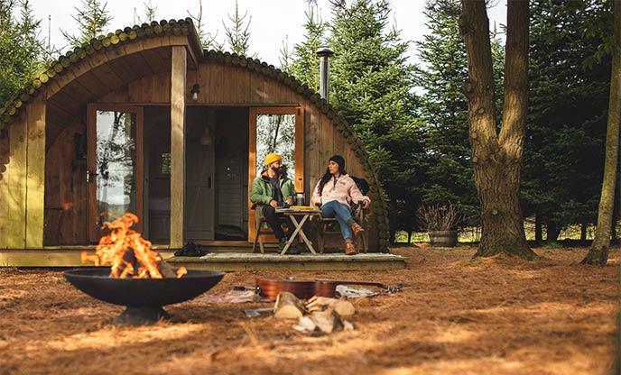 A couple sat outside a woodland cabin in the forest with a firepit
