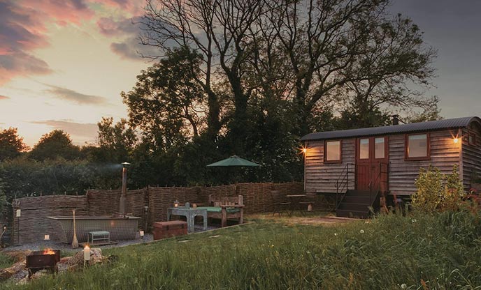 A view of the sun setting over Len's Hut in Somerset. The shepherd's hut and its outdoor bathtub are in the foreground, with trees behind them.